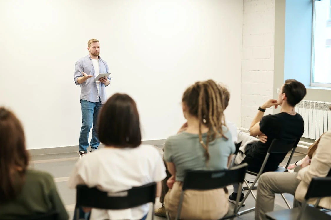 A picture of a man presenting in front of a small audience listening attentively